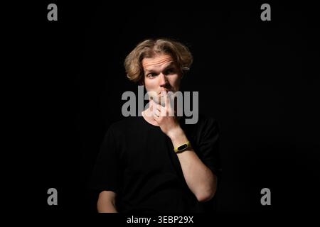 Young adult man posing with finger over lips expressing silence, secrecy, questioning, or deep contemplation on a dark background Stock Photo