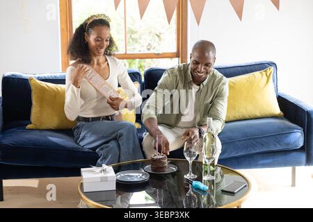 Couple with African American man and woman with sash sitting on navy sofa cutting cake Stock Photo