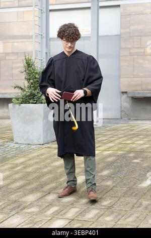 Man in twenties wearing black gown holding mortarboard with tassel and burgundy cover in courtyard Stock Photo
