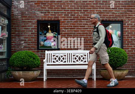 A senior male tourist with a backpack walks past a brick storefront and a white bench at Ghirardelli Square in San Francisco. Stock Photo