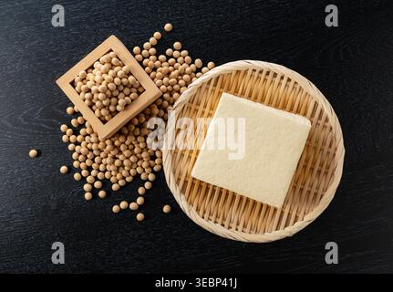 Block of firm tofu on a bamboo strainer with dried soybeans in a wooden measuring cup on a wooden table Stock Photo