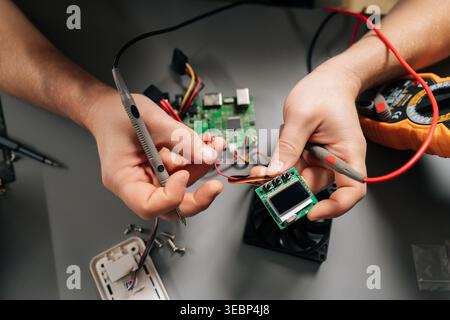 Close-up of repairman working with multimeter, testing circuits on display board connected to larger motherboard, surrounded by hard disk drive, fan Stock Photo