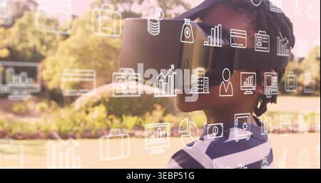 Standing boy wearing VR headset, visor cap, striped shirt in sunny park, displaying UI icons Stock Photo