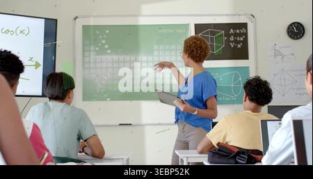 Pointing female instructor in blue blouse holding tablet, teaching in classroom with periodic table Stock Photo