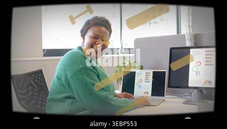 Smiling woman wearing chunky green sweater, headband, using laptop at desk with monitor and chart Stock Photo