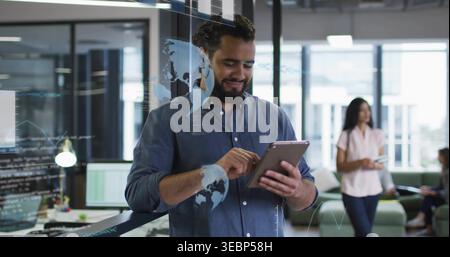 Leaning bearded man in blue shirt using tablet at open-plan office with glass partition overlays Stock Photo
