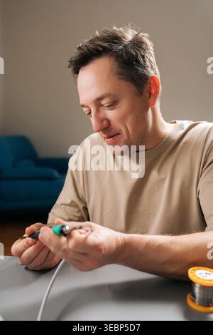 Cheerful young man soldering wires for repair, fixing cable connection with soldering iron, engaging in electronics hobby, preparing components for Stock Photo
