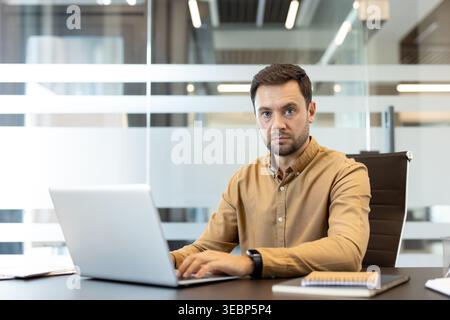 Man focusing on his laptop in a modern corporate office, looking directly at the camera with a serious expression while engaged in daily professional tasks and online communication Stock Photo
