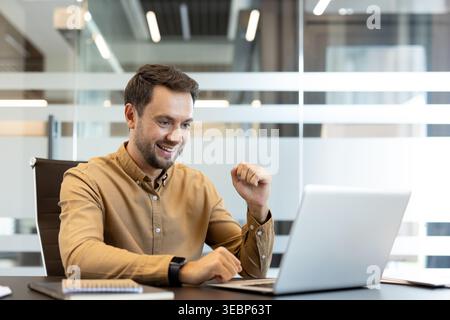 Young businessman looking at laptop screen, feeling excited and performing a winning gesture, celebrating business triumph or professional achievement in an office environment Stock Photo