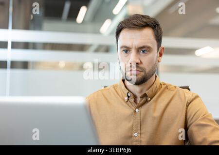 Serious young man concentrating on his computer screen while performing important tasks in a contemporary office environment, embodying dedication and professional focus Stock Photo