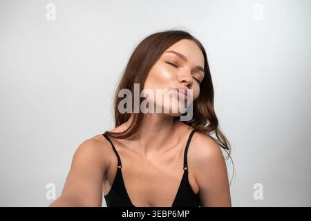 Young woman posing for a selfie, blowing a kiss with pouting lips and closed eyes against a clean white background Stock Photo