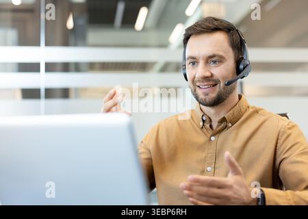 Man wearing a headset and brown shirt, communicating with a client through his laptop screen, actively gesturing and smiling while working in a modern office environment Stock Photo