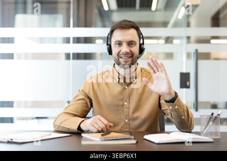 Man wearing a headset and brown shirt smiling, waving hello to the camera, and engaging in online communication with clients or colleagues while sitting at an office desk Stock Photo