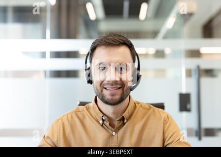 Friendly male customer service representative with headset smiling at camera in modern office, professional support agent ready to assist via phone, video or online chat Stock Photo