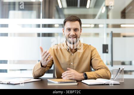 Businessman in a brown shirt at his desk, gesturing and speaking into a webcam during a virtual meeting, portraying hybrid work, remote collaboration and professional engagement Stock Photo