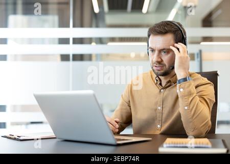 Man wearing a headset and brown shirt focusing on his laptop screen, actively engaged in a video call or customer service conversation while working in a contemporary office setting Stock Photo