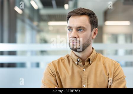 Serious young man contemplating or solving a problem, his intense expression and beard highlighting deep thought against a modern corporate background, representing focus and strategy Stock Photo