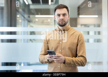 Serious male professional standing in a modern office using a smartphone, looking directly at the camera, communicating technology and business connection concepts Stock Photo