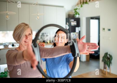 Senior woman performing physiotherapy exercise with ring under physiotherapist supervision at home. Stock Photo