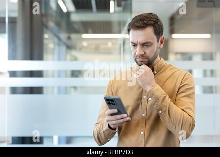 Man holding a smartphone, touching his chin, and looking at the screen with a serious and pensive expression, reflecting on information in a contemporary office setting Stock Photo
