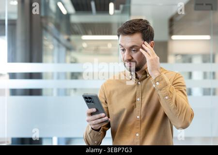 Young man experiencing surprise and disappointment while reading upsetting news or a shocking message on his mobile phone, showing a worried and emotional expression in a modern office environment Stock Photo