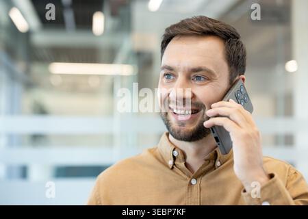 Young businessman happily communicating during a phone call, connecting with clients or colleagues in a bright professional workspace, representing corporate technology and success Stock Photo