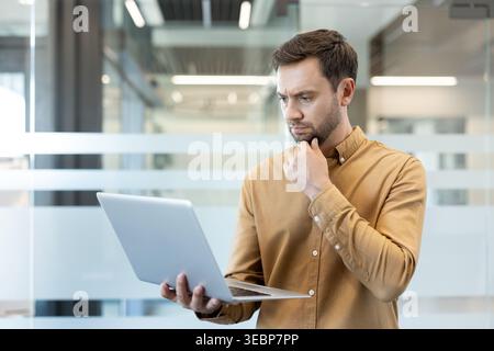 Thoughtful businessman standing in a modern office, concentrating while reviewing information on a laptop, contemplating solutions for business challenges and making strategic decisions Stock Photo