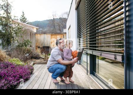 Father controlling smart home exterior blinds using tablet while standing with toddler son. Stock Photo