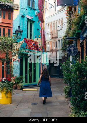 View of a woman in a blue dress walks down a vibrant, narrow alleyway with colorful buildings and hanging plants, a burst of urban artistry, London, U Stock Photo