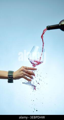 Dynamic splash of red wine pouring into tilted glass with hand on blue background Stock Photo