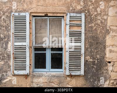 Rustic weathered wooden shutters on an old blue window frame set against a cracked stone wall of a traditional Mediterranean house in sunlight Stock Photo