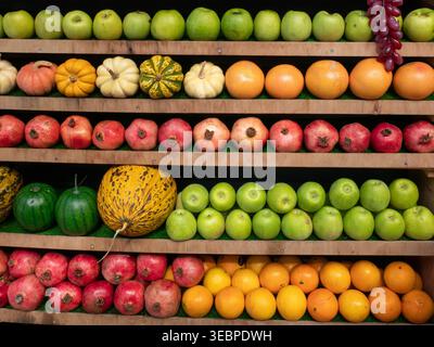 Vibrant organic harvest display with rows of fresh green apples pumpkins pomegranates oranges and seasonal melons on wooden market stall shelves Stock Photo
