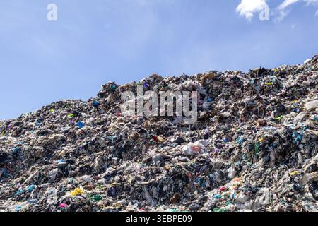 Large landfill site contains a high pile of various types of waste materials with visible plastic and organic debris under a clear sky during the day. Stock Photo