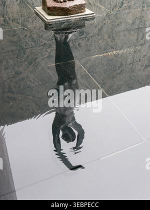 Striking reflection of a dark bronze statue of a diving female figure in a calm pool of water over tiled marble floor creating a moody scene Stock Photo
