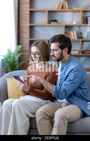 Couple sitting on a couch at home, looking at a smartphone screen, discussing content and pointing, engaged in online shopping and sharing digital con Stock Photo