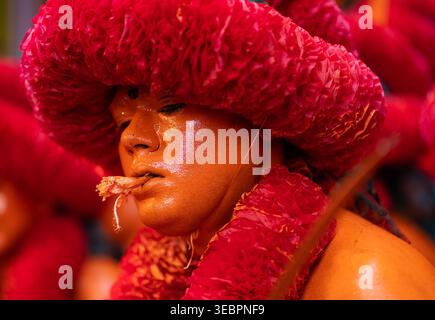 Munshiganj, Bangladesh - 13 April 2025: View of a performer in vibrant red headwear and ochre-painted skin, holding a fibrous object in their mouth. Stock Photo