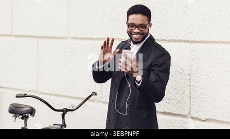 Man Smiling and Waving While Using Smartphone in Urban Setting With Bicycle Nearby Stock Photo