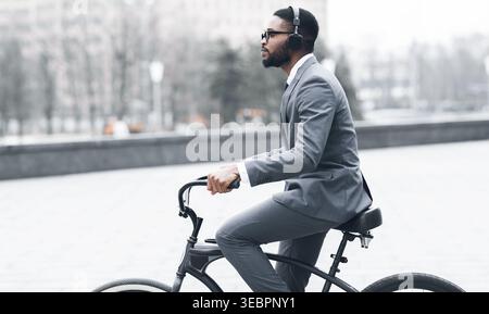 Man Rides Bicycle in City Wearing Suit and Headphones During Daytime With Urban Backdrop Stock Photo