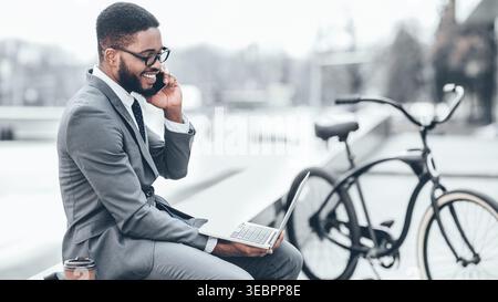 Businessman Using Laptop and Talking on Phone While Sitting Near Bicycle in Urban Setting Stock Photo