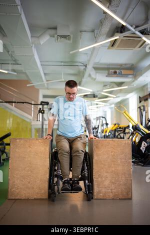 Confident man athlete in wheelchair doing push-ups strengthening exercise on wooden cubes in gym. Stock Photo