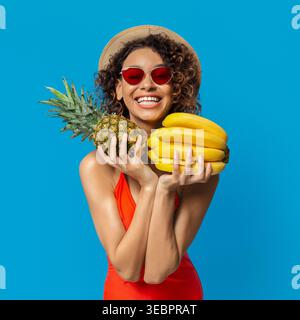 A Young Woman With Curly Hair and Red Sunglasses Smiles Widely While Holding Pineapples and Bananas Stock Photo