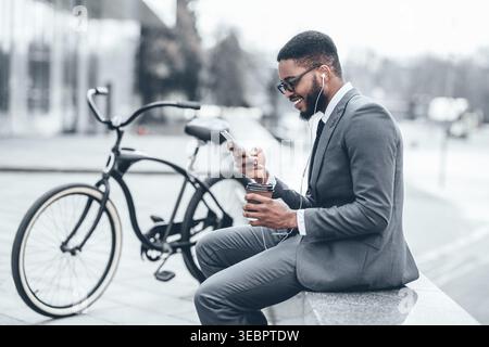 Man in Suit Enjoying Coffee and Browsing on Smartphone Near Bicycle in Urban Setting Stock Photo