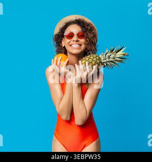 Smiling Woman in Swimsuit Holding Pineapple and Orange Against Bright Blue Background Stock Photo