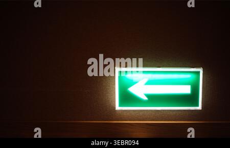Green illuminated sign with a left-pointing arrow on a dark wall, providing directional guidance in a dimly lit space Stock Photo