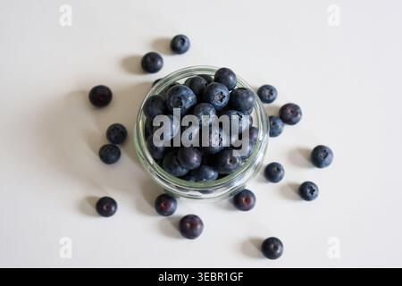 Fresh ripe blueberries in a small glass jar and scattered on a white background. Minimalist healthy food scene for summer and nutrition themes. Stock Photo