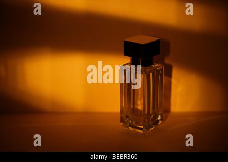 Glass perfume bottle with a black cap is highlighted by warm golden light, creating soft shadows on a smooth surface in a minimalist environment Stock Photo