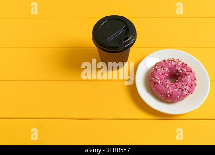 Pink Donut with Sprinkles and Black Coffee Cup on Yellow Wooden Background Stock Photo