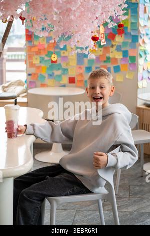 A joyful child in a light hoodie laughs at a modern cafe table, holding a plastic cup with a layered beverage. Background features pink cherry blossom Stock Photo