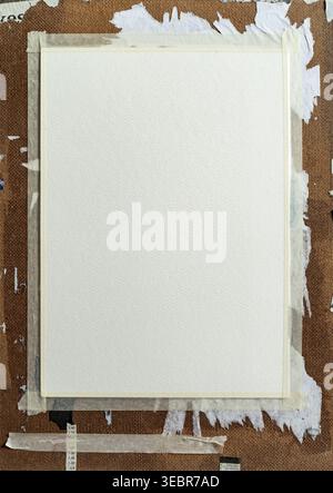 A vertical shot of a pristine sheet of cold-press watercolor paper secured to a distressed brown drawing board. The paper is framed with cream-colored Stock Photo