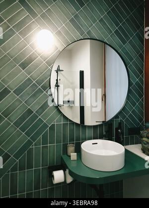 Bathroom interior with green diagonal tiles & round mirror. Modern bathroom with emerald green tiles in diagonal pattern, circular mirror, white sink. Stock Photo